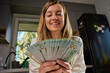 © Lazy_Bear - Smiling woman in kitchen counting cash while sitting at table with receipts and documents, managing home finances. Concept of planning home budget