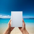 © SandraSevJarocka - People hands holding a white book mock-up with copy space in front of a beautiful beach