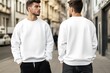 © Aleksander - Two young men wearing white sweatshirts mockup, standing back to back on a city street. The scene captures a modern urban environment with buildings and parked cars in the background.