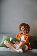 Wall Mural - Young joyful woman reading a book on a couch in a modern living room with a gray wall, wearing an orange shirt and white shorts, warm color palette