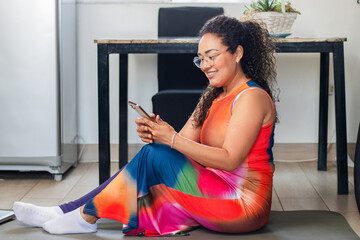 Middle aged brunette latin woman smiling sitting on the floor at home, using smartphone