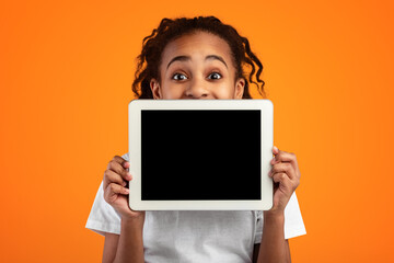 Great Application. Excited Black Girl Holding And Showing Digital Tablet With Empty Screen For Mockup, Covering Face And Mouth, Recommending New App Posing Standing Over Yellow Studio Background