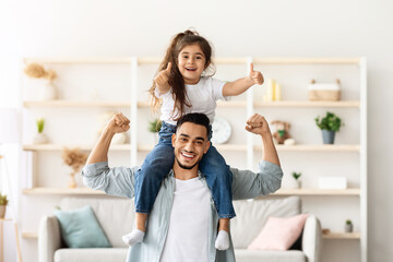 Wall Mural - Playful middle-eastern father and daughter having fun at home, cheerfully posing at camera, copy space. Happy arab man dad carrying little girl kid on his shoulders and showing strong muscles