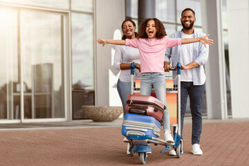 Wall Mural - Family Trip Concept. Portrait of cheerful African American girl having fun and spreading hands, ready for vacation, standing on luggage cart. Parents walking with baggage trolley, riding daughter