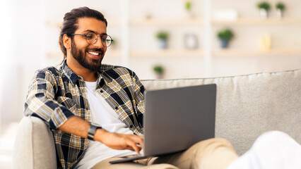 Happy indian guy with glasses working from home, using modern notebook, sitting on couch in cozy living room, copy space. Smiling middle-eastern man freelancer programming or chatting with ladies