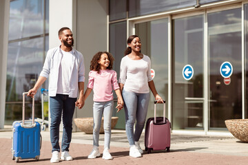 Wall Mural - Happy loving black family travelling together, walking with luggage suitcases near airport terminal building, outdoor area. Cheerful African American parents holding hands with daughter and smiling