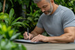 © Mselected - Man writing on tablet in a lush indoor garden setting with vibrant greenery