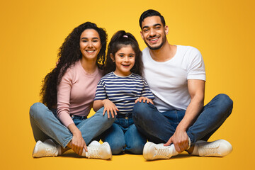 Wall Mural - Portrait Of Happy Young Middle Eastern Family Of Three With Little Daughter. Cheerful Arab Parents And Their Cute Female Kid Sitting On Floor Over Yellow Studio Background And Smiling At Camera