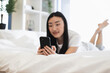 © sofiko14 - Asian woman in twenties lying on bed using smartphone. Relaxed atmosphere, connected with digital world, enjoying leisure time. Bright room, peaceful setting, modern lifestyle conveyed.