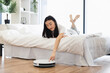 © sofiko14 - Asian woman relaxing on bed while controlling robot vacuum cleaner. Concept of modern technology, convenience, smart home lifestyle. Young adult enjoying clean and tidy living space.