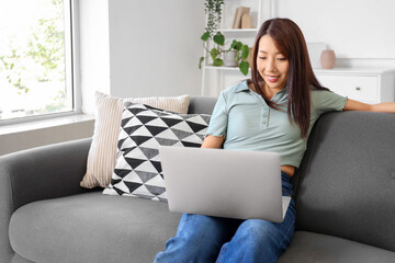 Wall Mural - Young Asian woman with laptop sitting on sofa at home