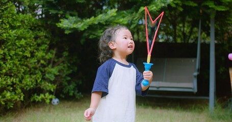 Wall Mural - A little Asian child enjoys Playing blowing Colorful soap bubbles Having Fun in a Lush Green Garden surrounded by greenery and sunlight on a Sunny Day, Joy Curiosity and Childhood Freedom Development