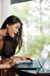 © SHOTPRIME STUDIO - Excited young woman with long dark hair using a laptop at a cozy workspace, surrounded by greenery, showcasing joy and enthusiasm for technology