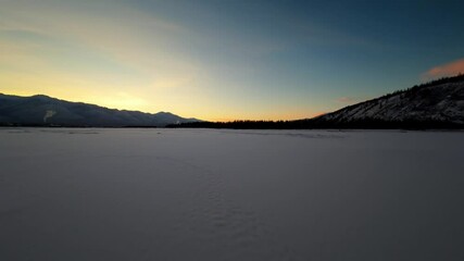 Wall Mural - A captivating sunset unfolds over a vast frozen lake, with majestic mountains and dense woods framing the tranquil winter scene. The sky transitions into warm hues as night approaches.