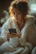 © Natalia - Early morning check-in: a person wrapped in a blanket, typing a quick update on a phone, coffee mug steaming close by,