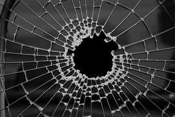  Black-and-white photograph of a shattered window, featuring a central void with spiderweb-like cracks extending outward. Jagged glass shards and intricate patterns evoke depth