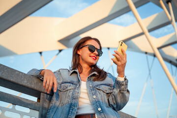 Wall Mural - Urban woman wearing sunglasses and denim jacket holding using mobile phone in front of city bridge.