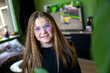 © Connect Images - Smiling young girl with glasses sitting indoors with a blurred background of a room with a computer monitor, Netherlands