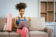 © NanSan - A young African American woman in workout clothes sits on the couch at home after a workout, smiling while looking at her computer, online shopping with a credit card in hand