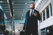 © BullRun - Contemplative corporate director with suitcase baggage and smartphone technology thoughtful looking away while searching terminal gate in airport, Caucasian employer 30s during business trip