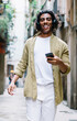 © BullRun - Smiling curly hipster guy in headphones walking on city street listening music and enjoying free time, positive young man meloman holding mobile phone with favorite songs in playlist using earphones