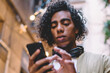 © BullRun - Pensive curly male holding mobile phone sending text messages in social networks chatting online, serious millennial hipster guy holding smartphone making banking via application standing on street