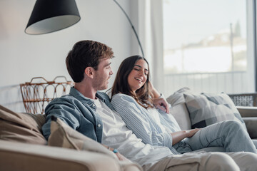 Wall Mural - Young happy couple having fun talking laughing relaxing at home on couch, boyfriend embracing girlfriend telling funny joke sitting on sofa, humor in relationships, enjoying weekend together.