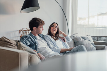 Wall Mural - Young happy couple having fun talking laughing relaxing at home on couch, boyfriend embracing girlfriend telling funny joke sitting on sofa, humor in relationships, enjoying weekend together.