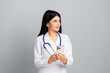 © deagreez - Young female doctor holding a smartphone in a professional white coat with stethoscope on a light grey background