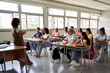 © Gigi Delgado - Diverse group of students attentively listening to their teacher in a bright classroom. The teacher stands at the front, engaging the class. The scene highlights education, learning and collaboration.