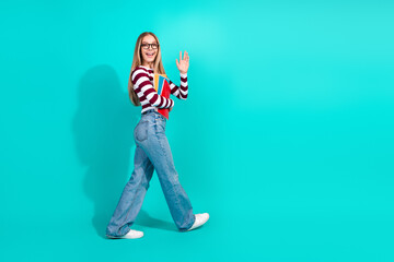 Happy student walking confidently with books against a vibrant turquoise background, stylish and youthful fashion