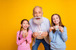 © deagreez - Cheerful grandfather and granddaughters enjoying together with tea cups against a vibrant yellow backdrop