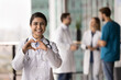 © fizkes - Indian female cardiologist in a white coat, showing heart shape with joined fingers, posing for camera in clinic office, embodying care, compassion, and dedication in healthcare services. Cardiology