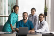 © fizkes - Four multinational clinic staff members, nurses and doctors posing, gathered at conference table with laptop, smiling, looking at camera with positive, joyful expression. GP portraits, success, career
