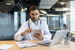 © Liubomir - Focused doctor sitting at desk with laptop. Man in white medical coat consulting patients remotely at workplace in office.