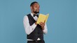 © DC Studio - Young doorman hotelier reading a book on his work break in studio, hotel concierge in tuxedo against blue background. Bellhop resort personnel enjoys literature and lecture. Camera B.