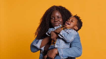 Wall Mural - Joyful african american mother carrying her adorable child on camera, holding him close and smiling in the studio. Happy mommy and cute little boy bonding together, loving parent. Camera A.