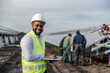 © Dusan Petkovic - Portrait of smiling interracial environmentalist typing on laptop at solar farm and monitoring works while workers installing solar panels in background.