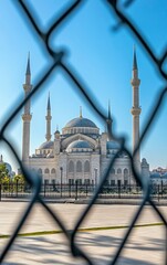 The white stone of the grand BWS shÃ—</ scored with intricate carvings and minarets, seen through black chain link fence in front, clear blue sky, mosque seen from far away, stock photo, 