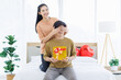 © Bangkok Click Studio - Portrait of young asian happy couple love surprise gift moment woman presenting man with box covering his eye with her palm sweethearts for Valentine's day in bedroom at home enjoy good time together