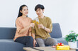 © Bangkok Click Studio - Asian young happy attractive couple love sweethearts smile drinking clinking holding champagne glasses sitting on cozy sofa in living room at home to celebrating anniversary enjoy good time together.