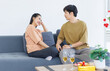 © Bangkok Click Studio - Asian young happy attractive couple love sweethearts smile hugging talking sitting on cozy sofa in living room at home to celebrating anniversary surprise enjoy good time together.