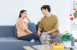 © Bangkok Click Studio - Asian young happy attractive couple love sweethearts smile hugging talking sitting on cozy sofa in living room at home to celebrating anniversary surprise enjoy good time together.