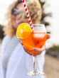 © simona - Close-up of a refreshing orange spritz cocktail with ice cubes and a straw, presented by a smiling woman, surrounded by a lively and festive summer setting ideal for aperitif gatherings
