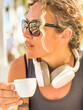 © simona - athletic woman relaxing outdoors in sunlight after a fitness session, wearing sporty casual clothes, sunglasses, white headphones, and holding an espresso coffee in her hand with a confident