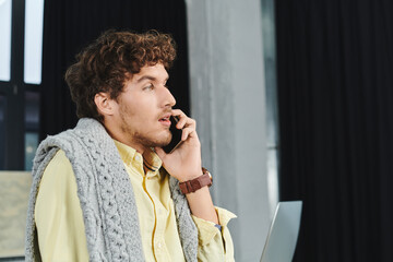 A young man with curly hair engages in a phone conversation while working in a stylish office.