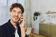 © LIGHTFIELD STUDIOS - Curly-haired young man smiles while talking on a phone in his modern, cozy apartment.