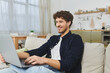 © LIGHTFIELD STUDIOS - A young man with curly hair smiles while working on his laptop in a cozy apartment.