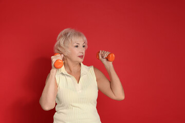 Senior woman exercising with dumbbells on red background, space for text