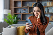 © Liubomir - Asian woman experiencing shock while reading phone message in home setting. Seated on sofa, she appears worried and surprised. Modern living room provides calm yet serious atmosphere.